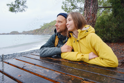 Couple enjoys Turkish coastline near Kemer. in pine forest. Sitting on stones, watching sea waves, relaxing together, slow romantic moment near calm blue water under soft winter light.