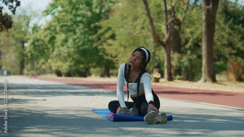 A young woman performs a fitness workout outdoors in a park. She focuses on arm exercises using a resistance band on a blue yoga mat.
