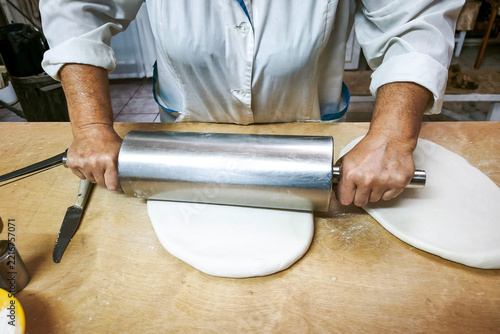 An elderly woman is rolling out dough with a large metal rolling pin.