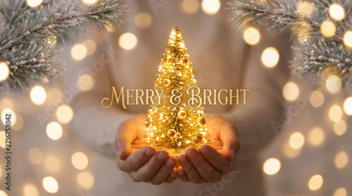 Hands holding glowing mini Christmas tree with Merry Bright text on bokeh background