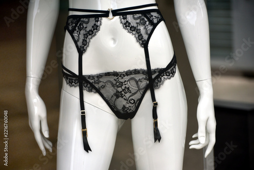 Close-up of black floral lace panties and a matching garter belt with adjustable straps and gold metal details, displayed on a white mannequin in a shop window.
