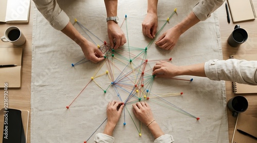 Several people s hands are working together to connect colorful strings on a beige cloth  surrounded by coffee cups and notebooks on a wooden surface