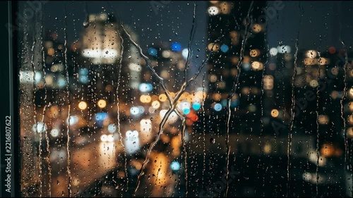 Rain drops are visible on a window. City lights blur in the background. The scene shows an urban setting at night with buildings lit up