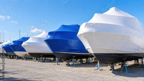 Row of boats in white and blue shrink wrap on stands in marina yard - winter boat storage