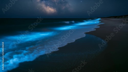 Bioluminescent waves roll onto dark beach beneath starry night sky. Glowing blue surf spreads across wet sand, creating serene, otherworldly coastal view with vivid luminous water.