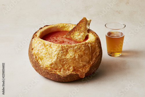 Grilled cheese rests in tomato soup inside a toasted bread bowl beside one small glass of amber beer