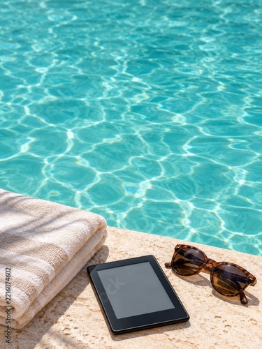 Electronic Reader and Sunglasses Beside a Towel at a Turquoise Swimming Pool