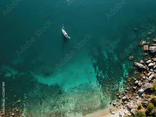 Aerial drone view of a white sailboat floating in turquoise tropical water near a rocky coastline