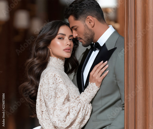 Couple in sage green tuxedo and cream lace blouse sharing romantic moment in warm wood paneled hallway.