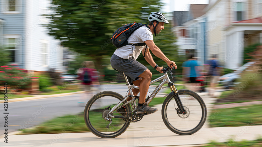 Fototapeta premium Man actively cycling a mountain bike on a city sidewalk with motion blur in a residential area, showcasing a healthy urban commute and dynamic outdoor exercise