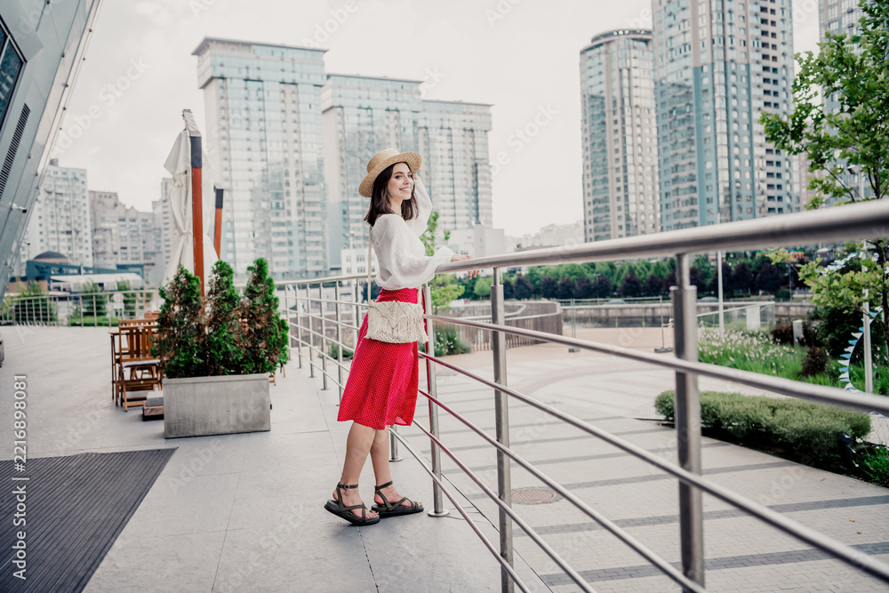 Fototapeta premium Young woman in a straw hat and red skirt enjoying a summer city waterfront view on an urban terrace