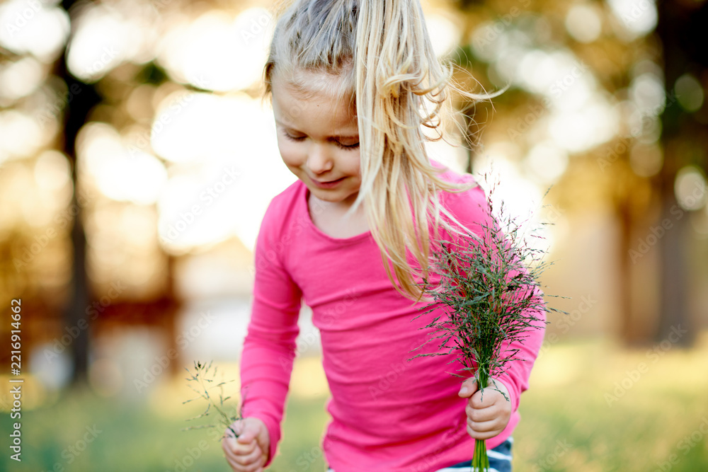 Fototapeta premium Park, child and pick grass to explore, play and development in environment on holiday. Smile, girl and kid gather plants in nature for collection, learning or growth on spring vacation with bokeh