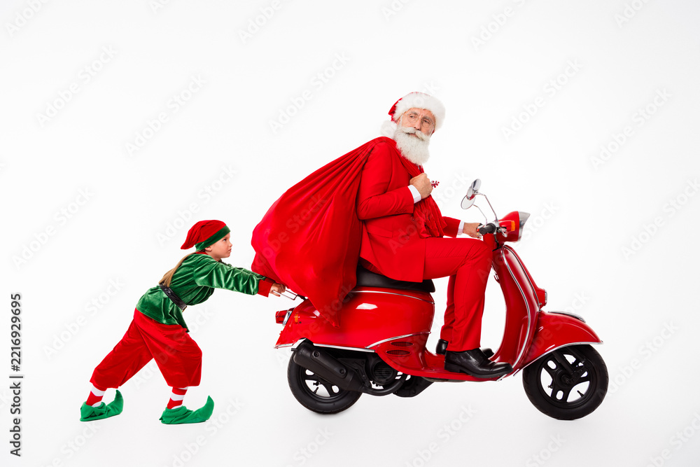 Fototapeta premium Santa Claus on a red scooter carrying a large gift sack while a playful elf child pushes in a whimsical Christmas holiday scene