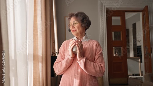 Senior Caucasian woman standing by window holding warm cup of tea and looking outside. Thoughtful elderly female enjoying peaceful morning coffee drink and quiet moment at home in sunlight.