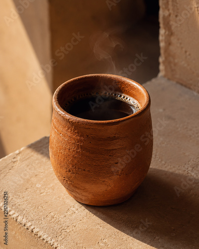Fresh hot black coffee served in a rustic handcrafted terracotta clay cup with steam rising on a warm stone ledge.