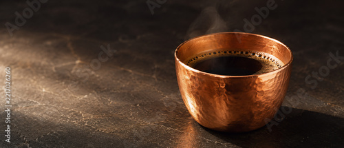 Fresh hot black coffee served in a rustic hammered copper cup with gentle steam on a dark textured marble surface.
