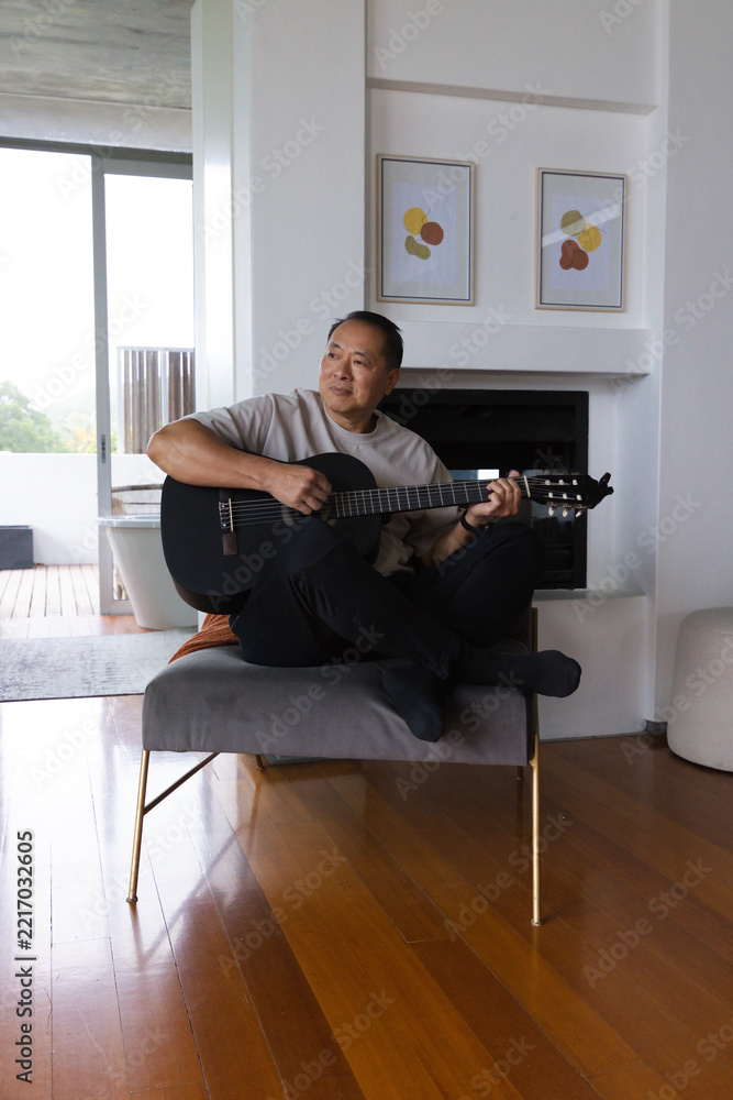 Fototapeta premium Senior Asian man sitting on bench playing black acoustic guitar in modern living room