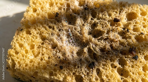 A macro close up view of a dirty yellow kitchen sponge covered in dark mold spots and white soap foam showing the porous texture of an unhygienic cleaning tool that needs replacement