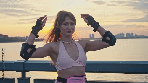 Beautiful girl fixes hair looking at camera at sunset. She stands on river promenade wearing protective gear enjoying warm summer lifestyle vibe after active rollerblading outdoor session.
