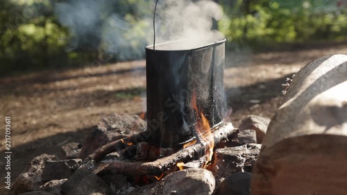 A black metal camping pot boils water over a stone-ringed wood campfire as thick steam rises from the top and flames flicker around burning logs in a slow motion outdoor close-up shot
