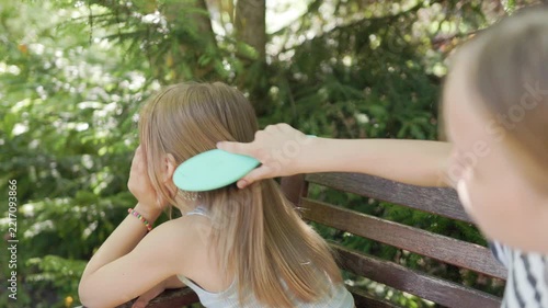 Gentle girl uses blue brush on straight blonde hair of sibling resting outdoors on sunny garden seat. Calm sisterhood moment reflects caring assistance, hygiene routine, and peaceful nature setting.