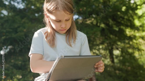 Cute schoolgirl operates touchscreen tablet while learning outside in nature park. Elementary student watches screen attentively, engaging with digital technology on sunny sunny afternoon.