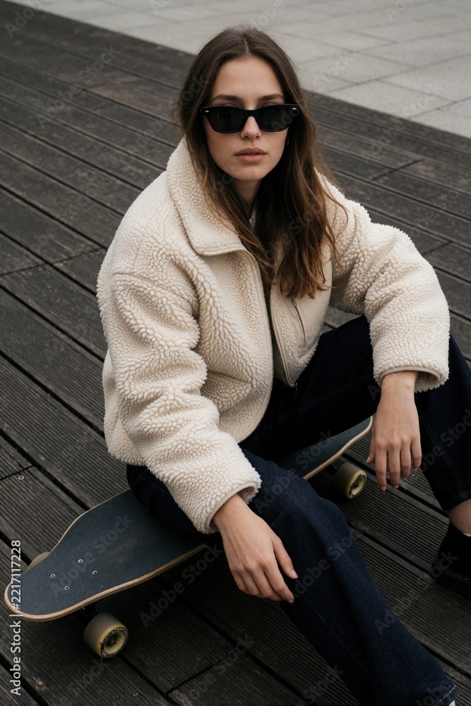 Fototapeta premium Young stylish woman in white fleece jacket, sunglasses sits on skateboard. Trendy girl relaxes on wooden pier boardwalk. Casual urban outfit with dark jeans, skater board for outdoor leisure activity
