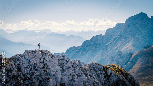 Hiker standing on a rocky summit during a via ferrata in the Austrian Alps near Innsbruck, Tyrol, with dramatic mountain peaks and a vast alpine landscape in the background.