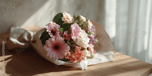A beautiful bouquet of assorted flowers, wrapped in paper and placed on a wooden surface. The soft sunlight casts gentle shadows across the scene