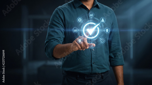 Torso of a person in a dark green shirt touching a glowing digital interface with a central checkmark icon surrounded by business and technology symbols