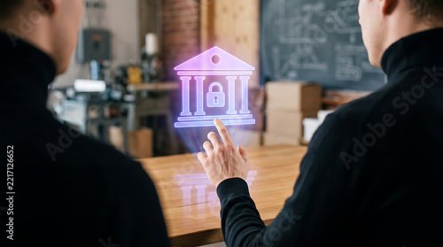 Two men in black sweaters looking at a glowing neon bank icon with a padlock symbol, projected above a wooden table in an industrial office setting