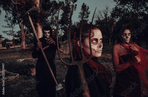 Three costumed figures with skull face paint holding pitchfork and scythe