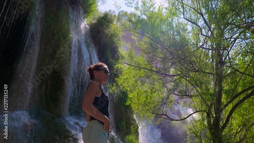Relaxed woman standing in front of a stunning waterfall, enjoying the refreshing spray and serene natural scenery during a sunny day