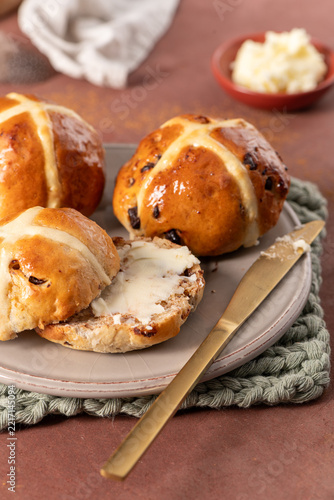 A close-up vertical shot of a sliced hot cross bun spread with melting butter on a grey plate with a gold knife in a rustic setting with cinnamon and Easter eggs