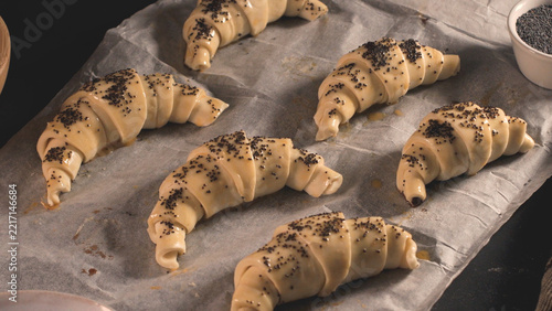 Raw unbaked croissants with poppy seed topping arranged on parchment paper on a kitchen countertop