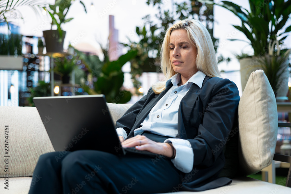 Fototapeta premium Focused businesswoman frowns while typing on a laptop from a cushioned rooftop terrace surrounded by plants, showing stress and frustration during flexible contemporary work outdoors.
