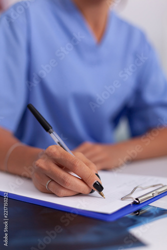 Close-up of a healthcare professional in blue scrubs writing notes on a clipboard, illustrating medical documentation, record keeping and clinical workflow.