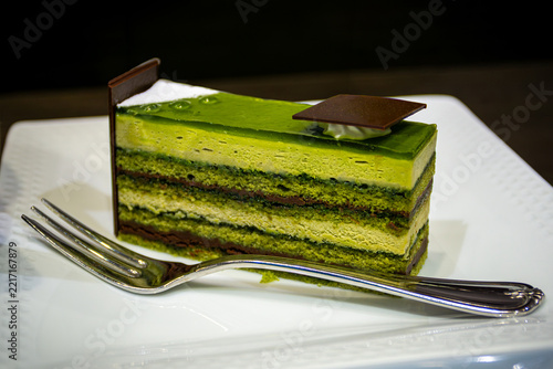 Closeup of a slice of Japanese matcha opera cake dessert with green tea layers of matcha sponge, cream, and chocolate ganache served on a plate with a fork.