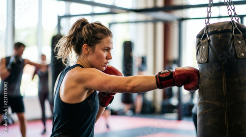 A determined woman with boxing gloves intensely punches a heavy bag in a busy, modern gym filled with other people training and exercising.