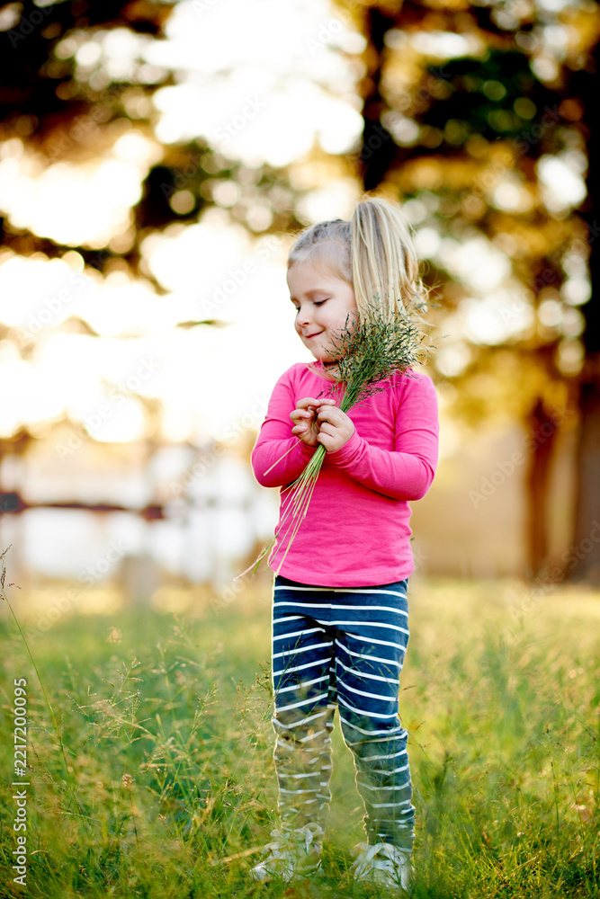 Fototapeta premium Nature, child and pick grass to explore, play and development in environment on holiday. Smile, girl and kid gather plants at park for collection, learning and growth on spring vacation with bokeh