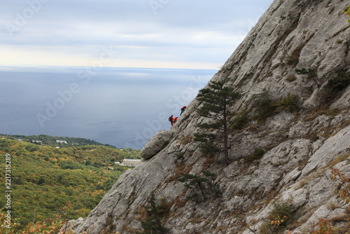 Rock climbers on mountain cliff in autumn forest, extreme ascent under cloudy sky