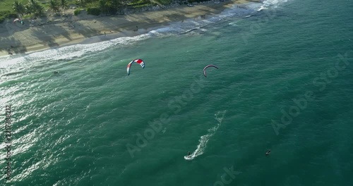 Two kitesurfers cross green water close to the Cabarete surfline under red and purple kites while curving white wakes appear beside the sandy beach and palms in the aerial view