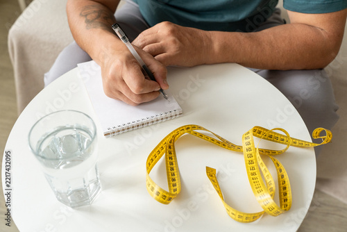 Man sits at table making notes in notebook, using measuring tape to track weight loss progress. He records health and weight data and monitors his metabolism