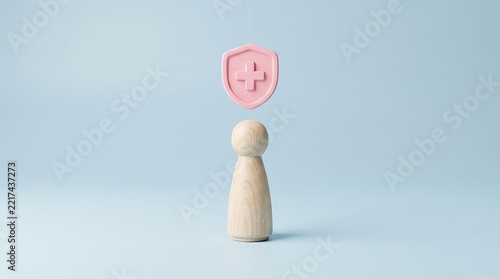Wooden figure figurine standing below suspended pink medical shield with plus sign symbolizing healthcare protection