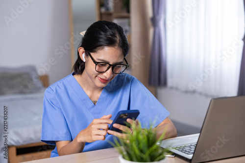 Asian nurse in blue uniform using smartphone beside laptop at home