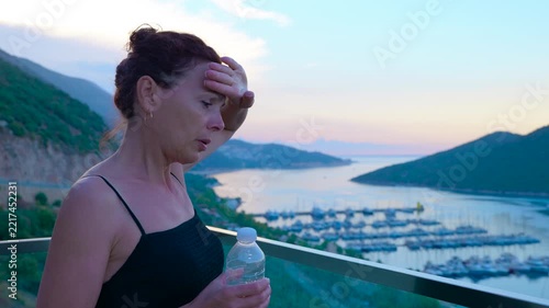 Woman drinking water on a balcony at sunset with a sea view. Woman feeling thirsty opening a bottle and drinking water while standing on a balcony overlooking a marina with boats during a sunset