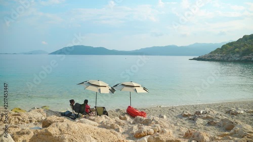 Couple relaxing on a secluded beach enjoying the seascape. Two people sitting under umbrellas on a remote rocky beach, admiring the calm turquoise sea and distant mountains during a peaceful vacation