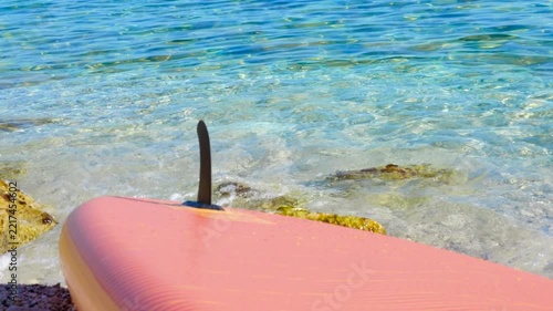 Pink stand-up paddleboard resting on tropical beach shore. Summer scenery showing a pink paddleboard on a sunny pebble beach with clear turquoise water gently lapping the shore