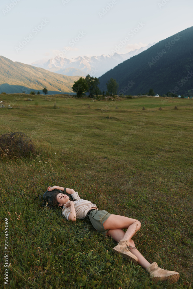 Naklejka premium Calm portrait of a woman resting in a meadow beneath distant hills, enjoying outdoor space, sunlit grass and tranquil scenery, a quiet moment of leisure and nature.