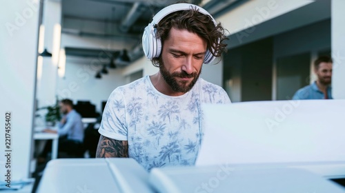 Young man with headphones concentrating at a large office printer in a modern coworking space